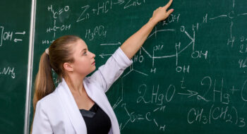 Woman standing in chemist's classroom near board and pointing on it